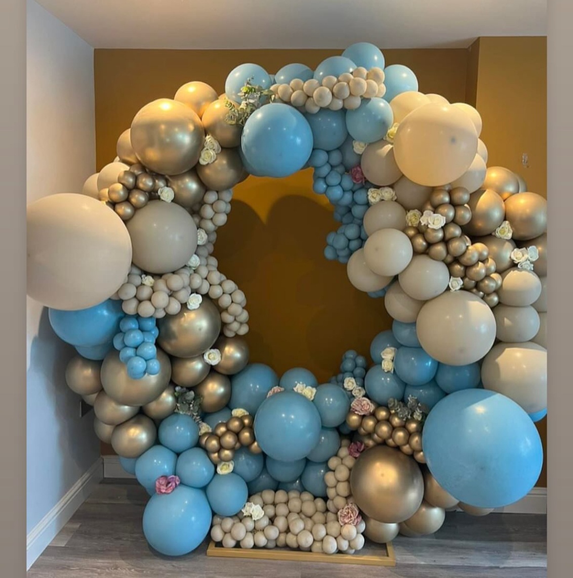 A large organic balloon Moongate in pale blue, cream and chrome gold, dotted with cream and blush roses, against a mustard wall.