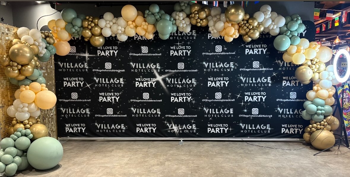Sage, peach, cream and gold balloon garland arching over the Village Hotel Club “We love to party” backdrop.