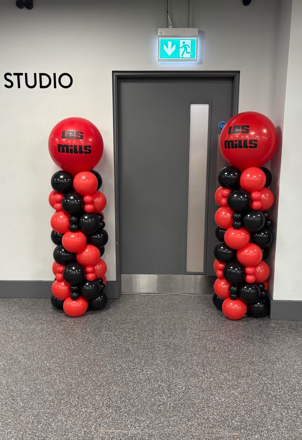 Two tall red and black balloon columns, each topped with a branded Les Mills balloon, either side of a fitness studio door.
