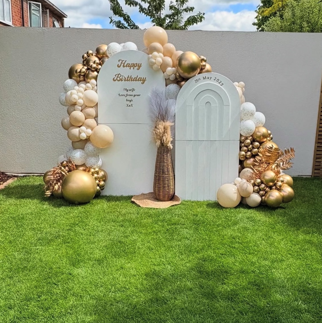A neutral sailboard display: two white arched boards with a Happy Birthday message, framed by cream, nude and gold balloons and pampas on a lawn.
