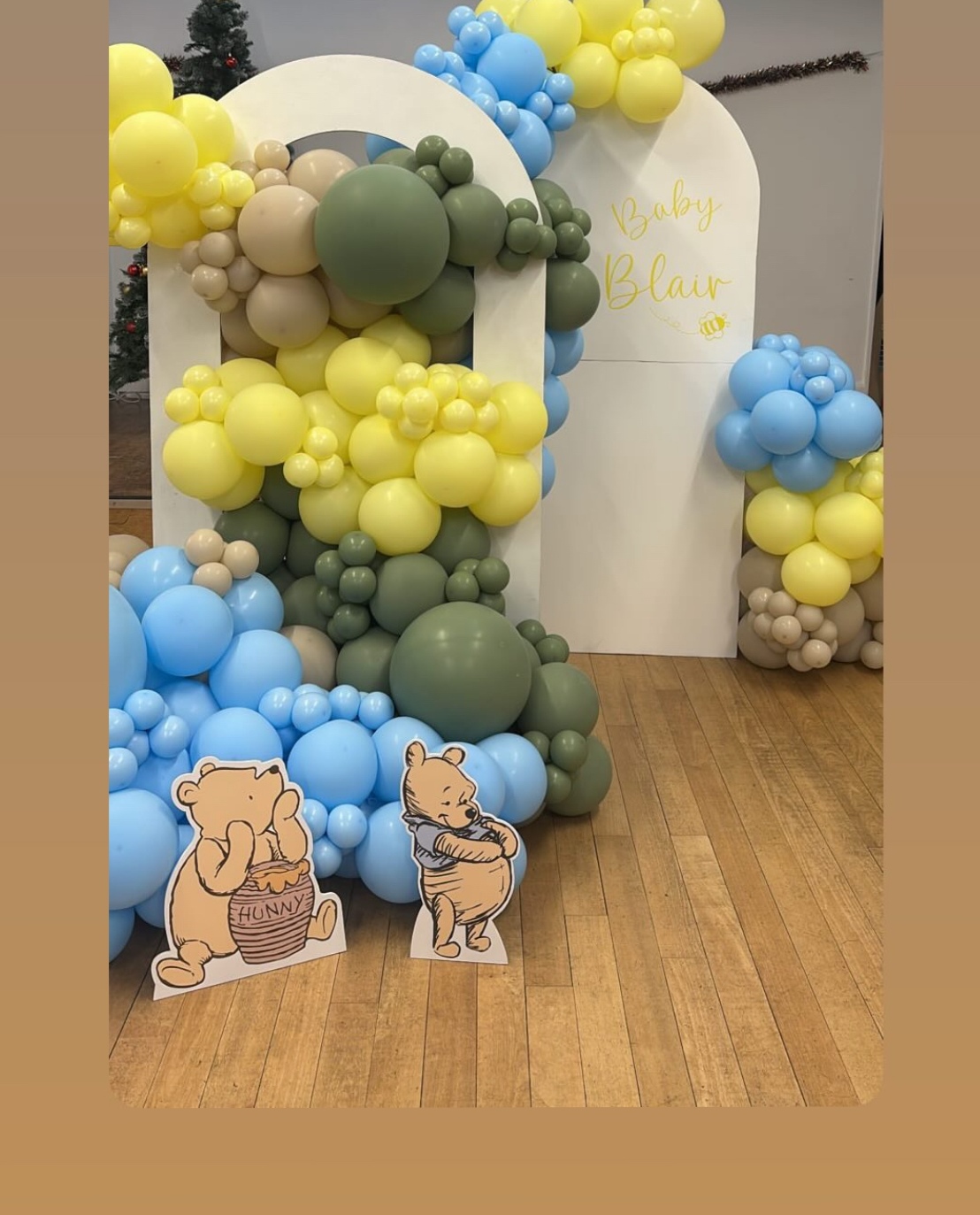 A Winnie the Pooh baby shower backdrop: arched boards and a yellow, sky blue and olive green balloon garland with Pooh and Piglet cut-outs.
