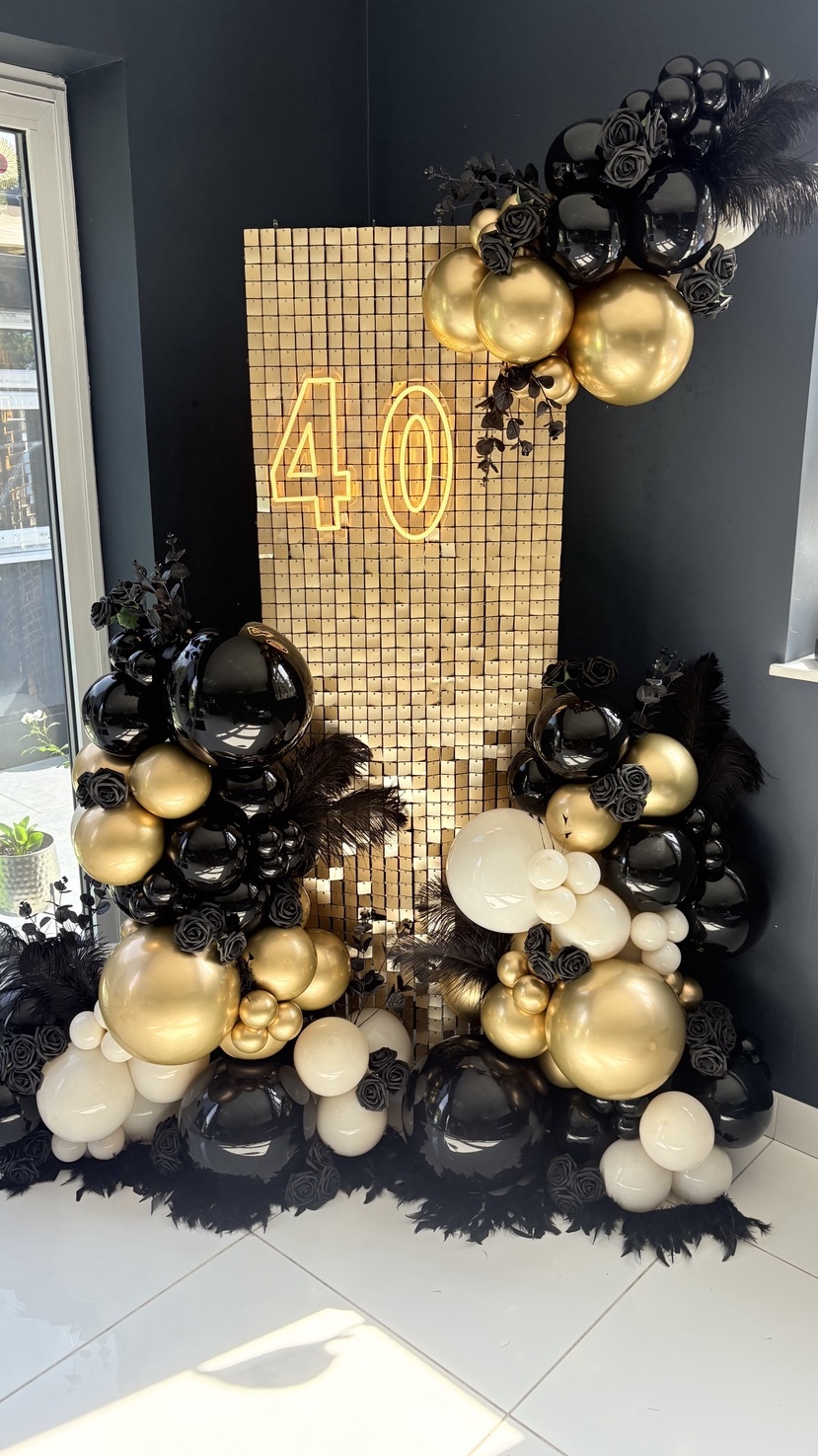 A 40th birthday display: a gold sequin panel with a neon 40, dressed with black, gold and white balloons and black feathers.
