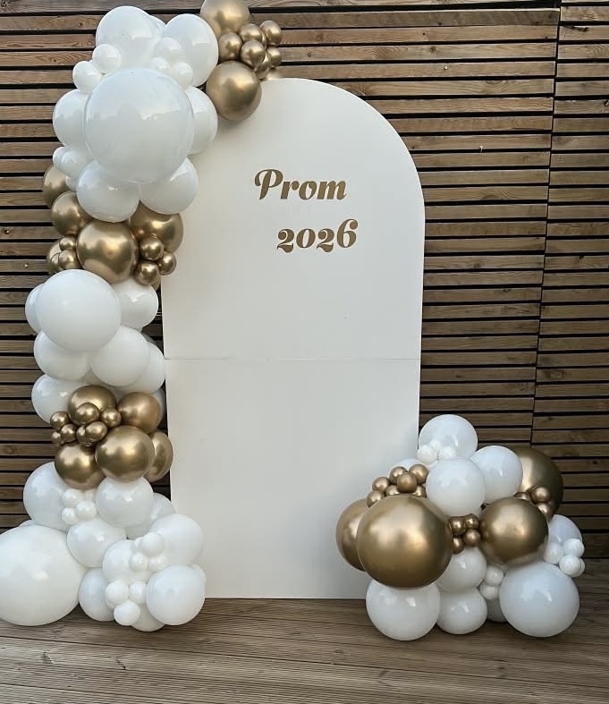 A white arched “Prom 2026” board framed with white and chrome gold balloons.
