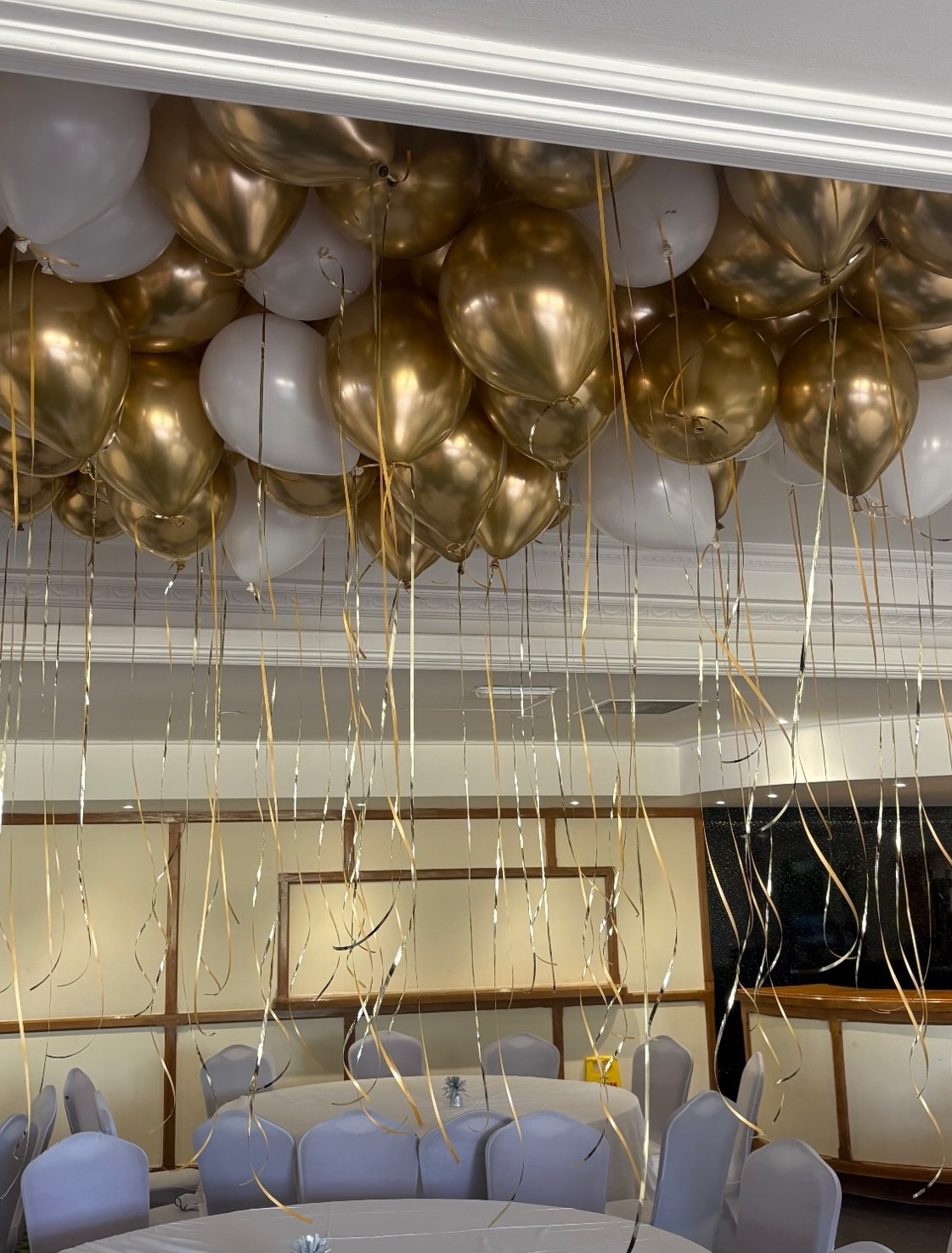 A ceiling of chrome gold and white helium balloons with gold ribbons trailing over a dressed function room.
