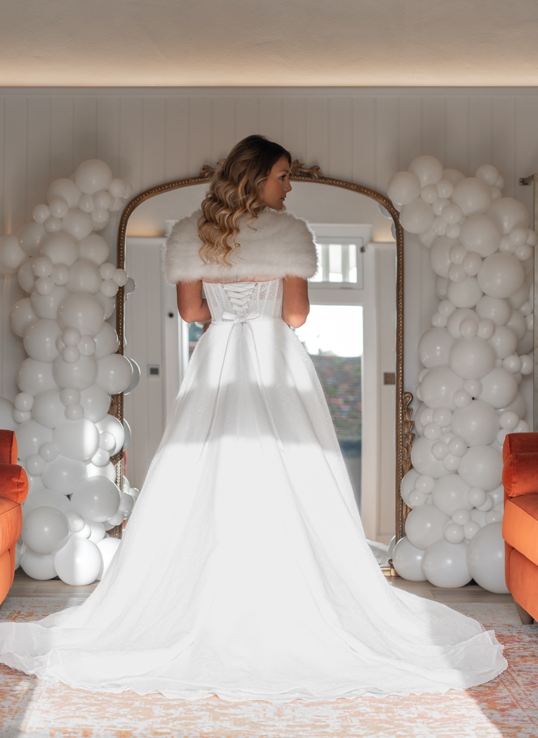 A bride in a white gown before an ornate mirror framed by an all-white organic balloon installation.